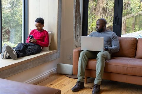 African American Father Working on Laptop While Son Uses Smartphone on Cozy Window Seat