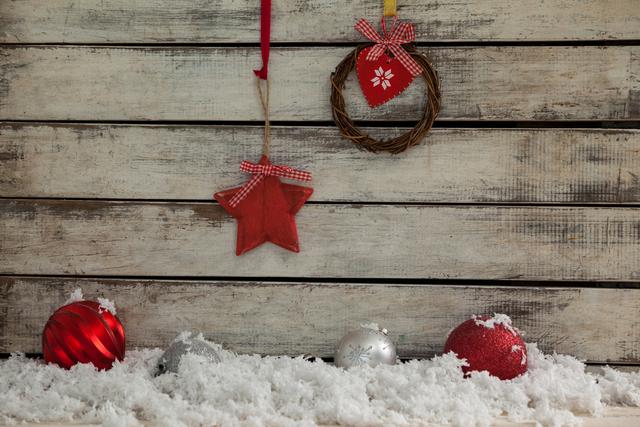 Rustic Christmas decorations featuring a wooden background with hanging wreath and star ornaments. Snow and colorful baubles add a festive touch. Ideal for holiday greeting cards, seasonal promotions, and festive home decor inspiration.
