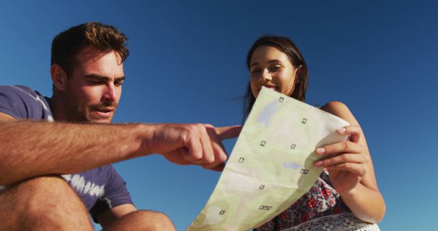 Young couple outdoors under clear blue sky, holding and navigating with a map. Ideal for concepts of travel, adventure, vacation planning, outdoor activities, teamwork, and exploration.
