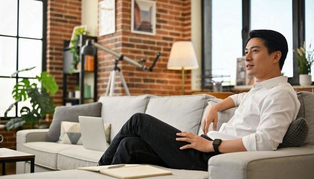 Relaxed Asian man working remotely on laptop in modern industrial loft living room