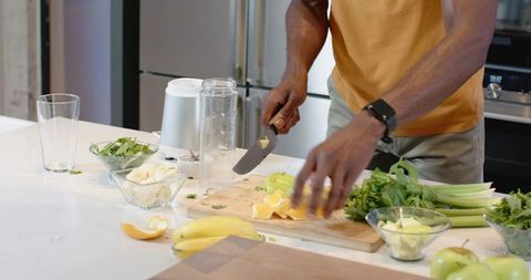 Man chopping citrus and greens on kitchen island preparing smoothie, wearing mustard tee
