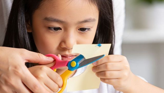 Asian girl cutting paper with child-safe scissors, adult guiding for craft learning, home