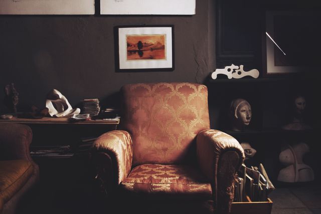 Depicts a vintage armchair in a dimly lit study room filled with books, art pieces, and sculptures. Creates a sense of nostalgia and comfort suitable for themes related to relaxation, classic interiors, or solitary moments. Ideal for use in lifestyle blogs, interior design portfolio, or vintage collection features.
