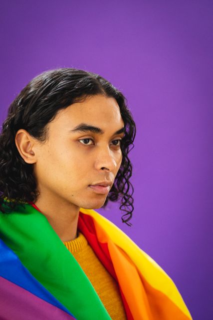 Close-up of biracial thoughtful young man with rainbow flag looking away against purple background. Copy space, unaltered, gay, contemplation, lgbtqia rights, lgbtqia culture and pride concept.