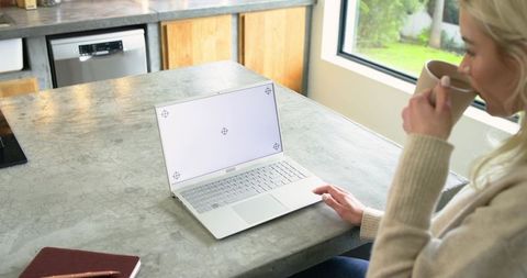 Woman Working Remotely at Kitchen Island, Drinking Coffee and Typing on White Laptop