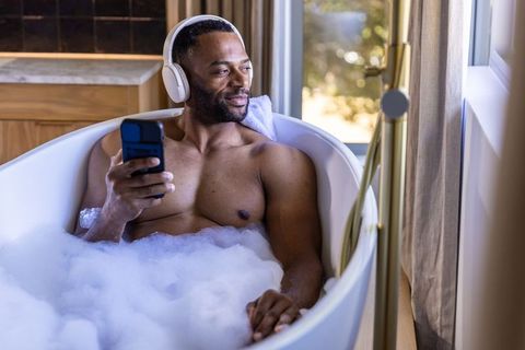 Mature black man relaxing in bubble bath listening to music with headphones and smartphone