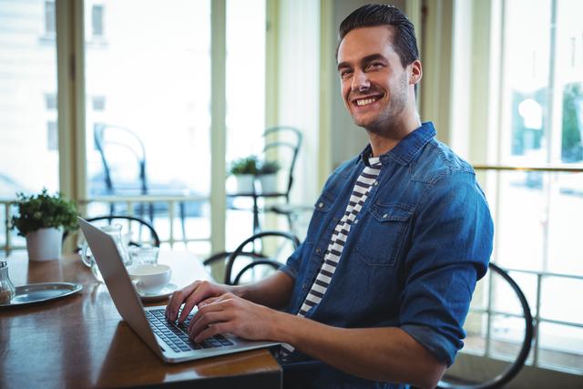 Young man in denim shirt using laptop in a modern café. Perfect for illustrating freelance work, digital nomad lifestyle, remote working, business environment, student life, or casual professional settings.