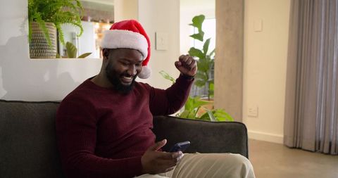 African American Man Wearing Santa Hat Celebrating on Smartphone in Cozy Living Room