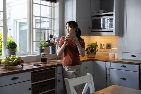Asian woman standing at kitchen window holding coffee mug french press morning routine