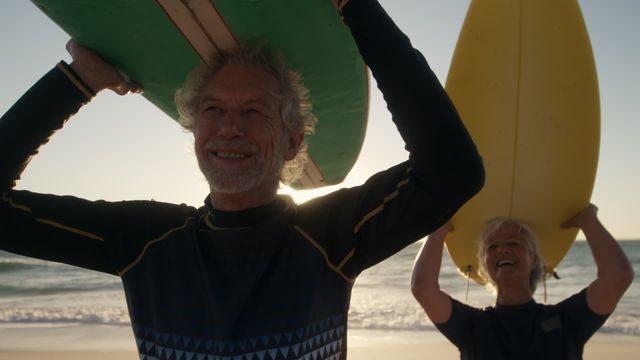 Senior couple carrying surfboards heads along beach at sunset, embodying active retirement and carefree lifestyle. Ideal use in promotional material for travel, retirement living, or wellness advertising focusing on joyful aging and outdoor adventures.