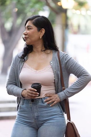 Young woman standing holding takeaway coffee cup on city sidewalk wearing cardigan and jeans