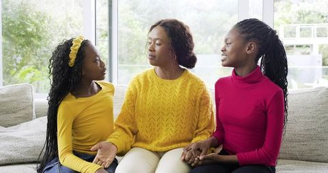 African American mom and daughters sitting on sofa holding hands and talking
