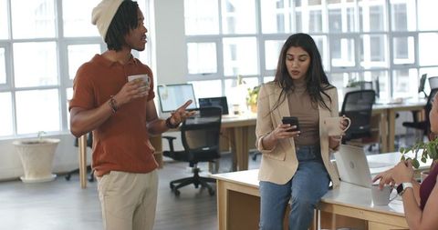 Diverse coworkers chatting in modern open-plan office woman using smartphone and coffee