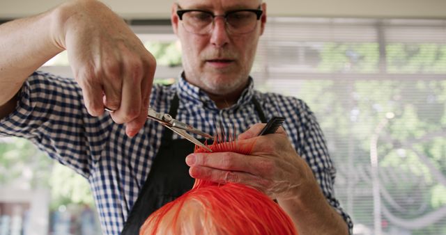 Senior hairdresser wearing glasses and apron, cutting orange hair with precision in modern salon. Ideal for use in articles on hairdressing techniques, hairstylist profiles, salon services promotion, and grooming essentials.
