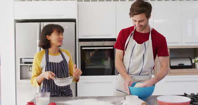 Young couple happily baking together in a modern kitchen. Both individuals are wearing aprons and are actively engaged in the baking process with various ingredients around them. Perfect for use in blogs, advertisements, or social media posts about home activities, cooking together, and family life.