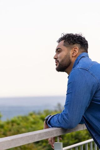 Contemplative man leaning on railing overlooking ocean horizon wearing denim shirt with smartwatch