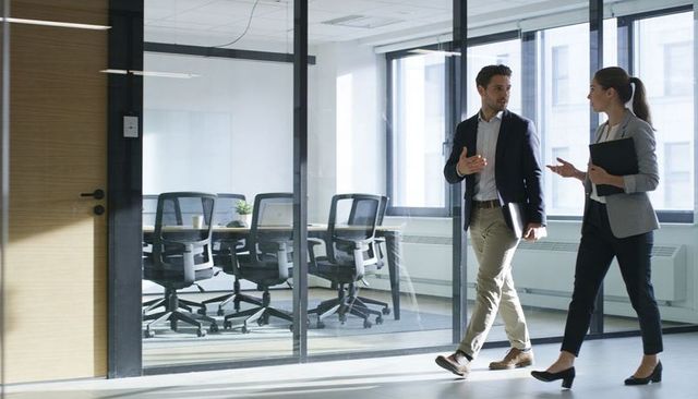 Walking business colleagues talking in modern glass office corridor holding laptop and folder