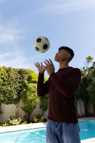African american teen tossing soccer ball by backyard pool on sunny day