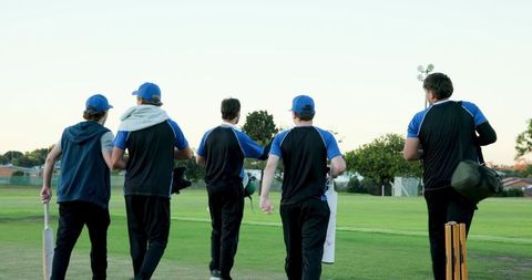 Five cricketers walking off pitch carrying bats and gear during evening cricket practice