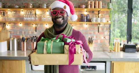 Middle-aged african american man smiling holding wrapped christmas gifts in kitchen with santa hat