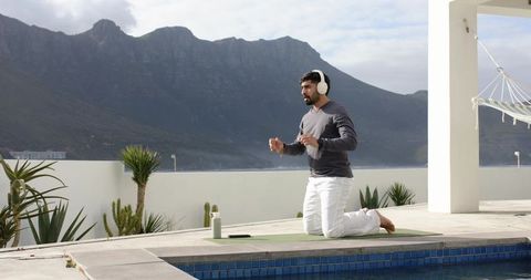 Indian man practicing yoga on rooftop pool deck wearing headphones with mountain coast view