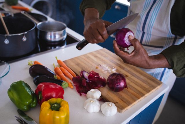 Senior African American man wearing apron chopping vegetables in a modern kitchen. Ideal for content related to healthy eating, active retirement lifestyle, home cooking, and culinary skills. Perfect for articles, blogs, and advertisements promoting healthy living and senior activities.