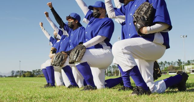 Diverse team of female baseball players in face masks kneeling in line with arms raised. female baseball team, sports training and game during coronavirus covid 19 pandemic.