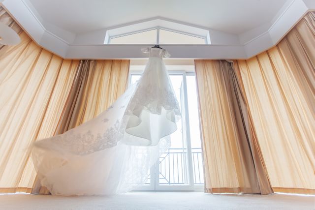 Bright image showing a lace wedding dress hanging elegantly in a well-lit room with sheer curtains. Perfect for bridal blogs, wedding planners, fashion catalogues, or interior design showcases.