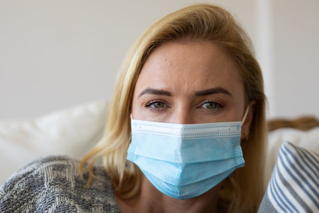 A caucasian woman sitting on the couch at home with a serious expression on her face. she is wearing a facemask