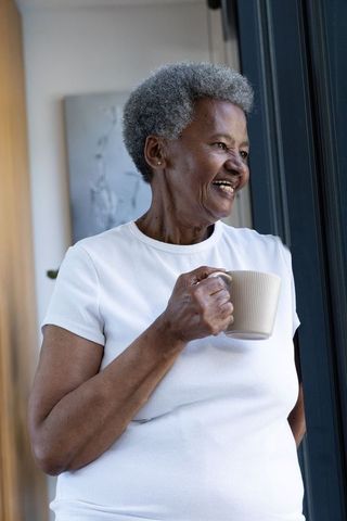 Senior african woman standing by window holding coffee mug smiling in sunlit home