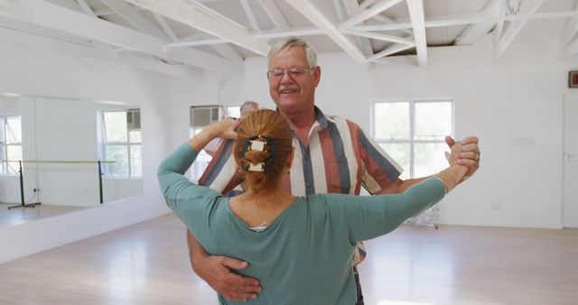 Elderly man and woman dancing together in a bright dance studio. The couple is facing each other with arms up, enjoying themselves. This image can be used for content related to senior fitness, healthy aging activities, couple bonding, or leisure activities for retired individuals.