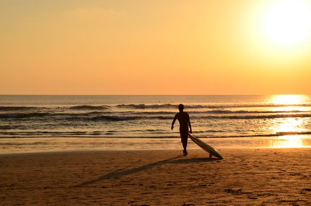 Surfer is walking along beach, carrying surfboard, with ocean waves and setting sun in background. This image evokes feelings of adventure, relaxation, and summer fun. Ideal for travel blogs, surfing promotional materials, and lifestyle articles focused on beach activities.