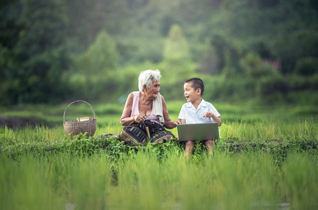 Image depicts an elderly woman and a young boy sitting in a green rice field, engaging with a laptop together. Ideal for illustrating themes of technology bridging generations, rural education, family bonding, and connections between tradition and modernity in rural or agricultural settings.