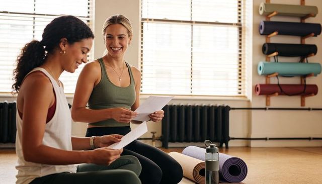 Two women reviewing yoga lesson plans in sunlit studio with rolled mats and water bottle