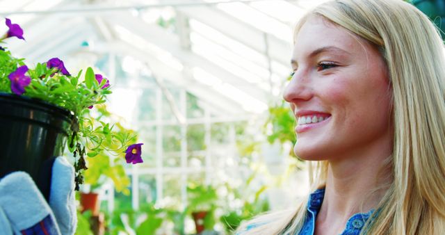 This vibrant image showcases a smiling blonde woman enjoying her gardening activity in a bright greenhouse. She is holding a flower pot, wearing gardening gloves, and surrounded by lush greenery and sunlight. Ideal for articles on gardening tips, horticulture guides, environmental blogs, and nature-related advertisements.