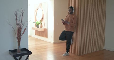 African american man leaning on wood slats using phone and headphones in sunlit hallway