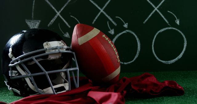 A football helmet and ball rest on a red cloth against a chalkboard with game strategy, with copy space. It captures the essence of American football planning and strategy development.