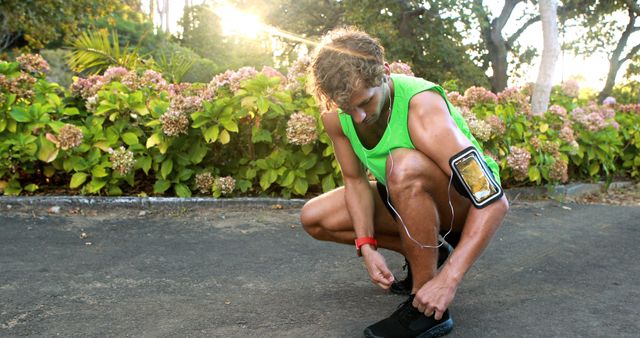 Male athlete is seen tying his shoelaces before a morning run in a park. He is dressed in a green tank top and uses an armband to hold his smartphone, demonstrating readiness for exercise. Trees and bushes bloom in the background, indicating a natural and vibrant setting. This image is ideal for promoting fitness, healthy lifestyle tips, exercise motivation, athletic wear, and outdoor activities.