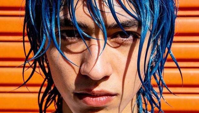 Staring young man with wet electric blue hair facing camera in sunlit orange metal shutter