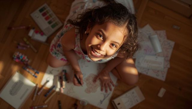 Smiling girl coloring on sunlit wooden floor wearing floral dress and using markers