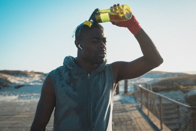 African American man taking a break from his workout on the beach at sunset, pouring water on himself to cool down. Ideal for promoting fitness, healthy lifestyle, outdoor activities, and hydration. Suitable for use in fitness blogs, health and wellness websites, sports advertisements, and summer activity promotions.