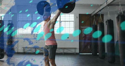 Fit man lifting barbell overhead in gym near punching bags with teal digital overlay