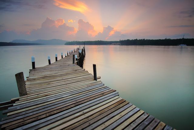 Tranquil wooden pier stretching out over a calm lake at sunrise with vivid rays breaking through clouds. Ideal for promoting relaxation, nature retreats, outdoor photography, and tranquility. Perfect for travel brochures, serenity-themed websites, meditation apps, and landscape-inspired decor.