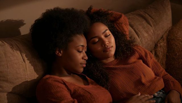 Two women cuddling on sofa in warm amber light, cozy intimate evening at home
