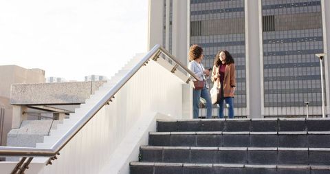 Two women talking on urban stairs with handrail and bags, sunlit candid street scene